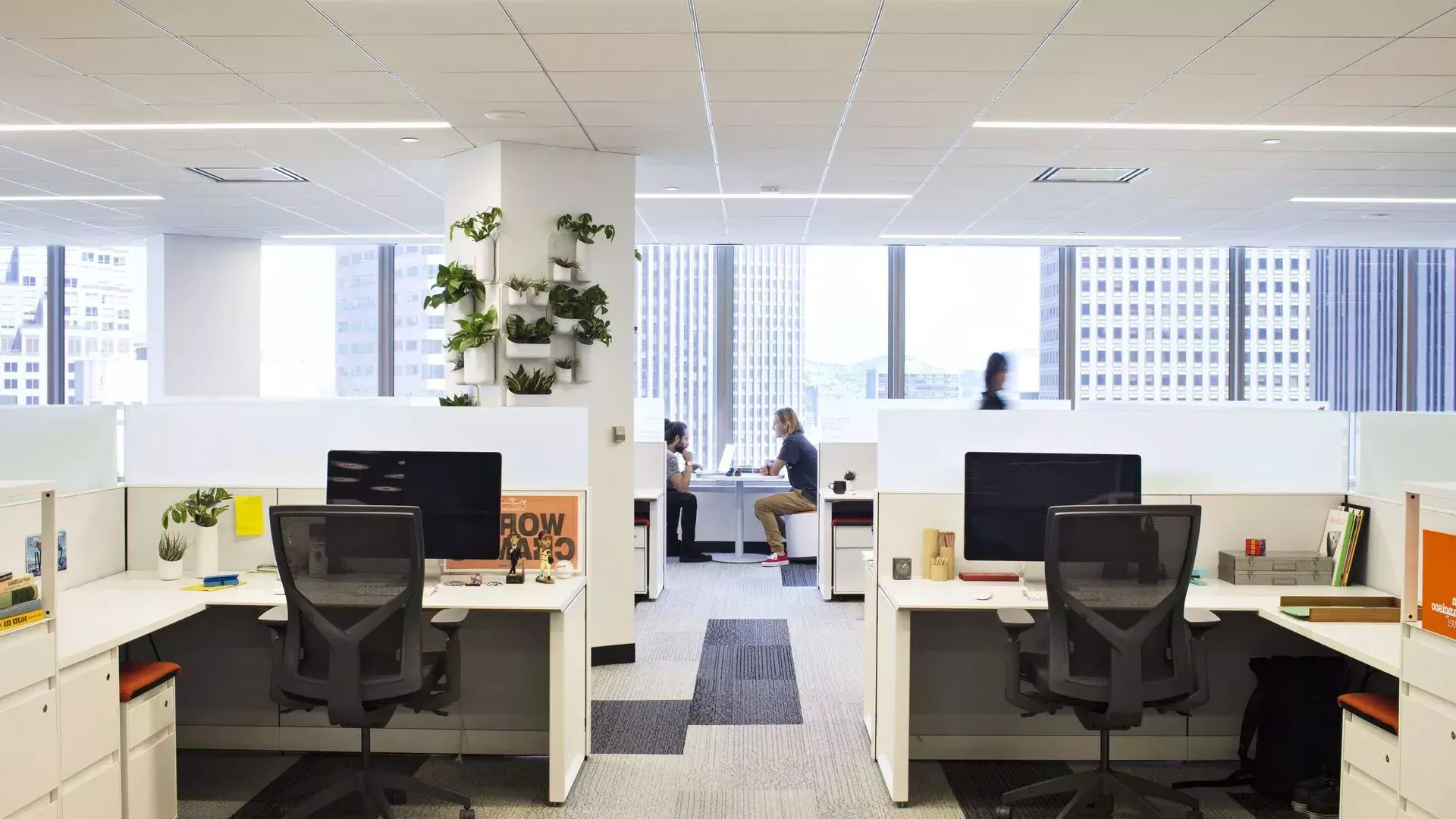 贝博体彩app一座办公大楼的室内场景, with desks in the foreground and two people working at a table in front of large windows.