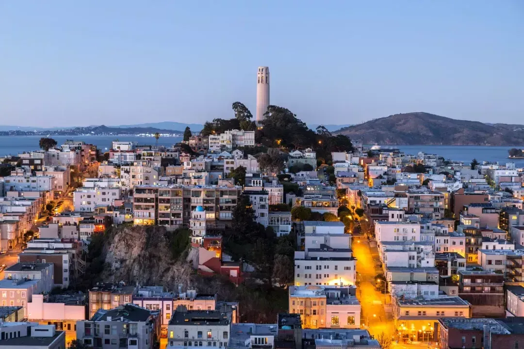 La Coit Tower de San Francisco au crépuscule, avec des rues éclairées devant elle et la baie de San Francisco derrière elle.